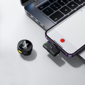 A small wireless Hoco microphone lying on a table next to a laptop and smartphone.